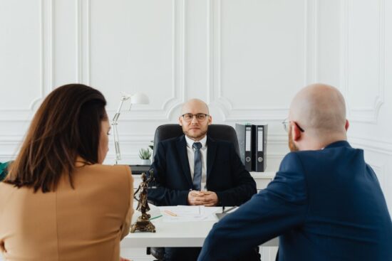 A bald man in a dark suit sits behind a white desk, facing two coworkers across the desk in a bright, white-paneled office setting.