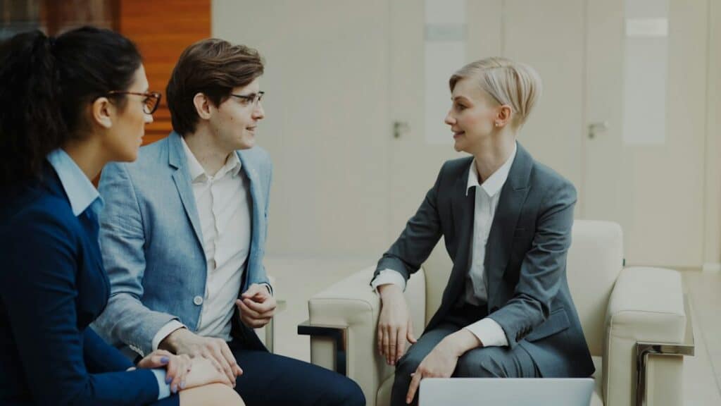Three professionals in business attire sit in a bright office lounge, chatting and smiling.