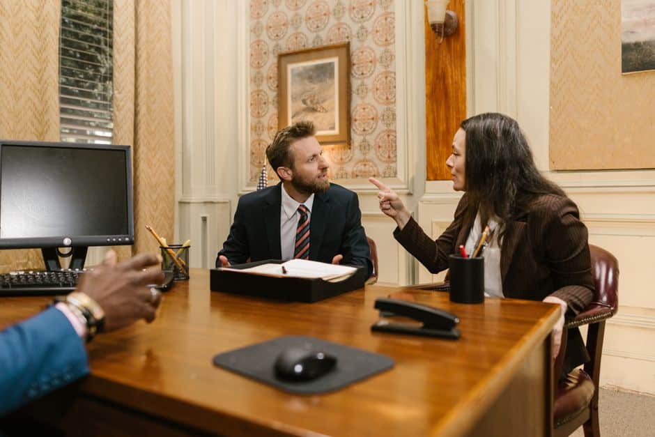 Two professionals discuss documents at a wooden desk in a formal, ornate office.