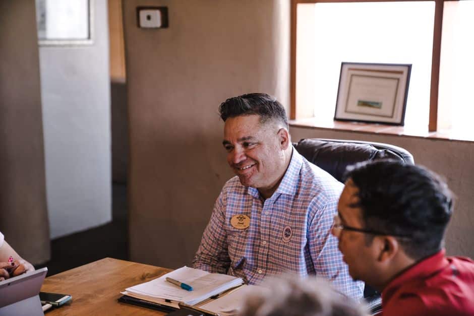 Smiling man in a checkered shirt seated at a table with papers and a name badge during a meeting in a cozy office.
