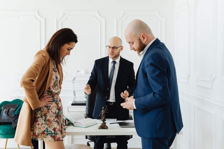Three professionals in a bright office discuss documents at a desk, one man gesturing as others listen.