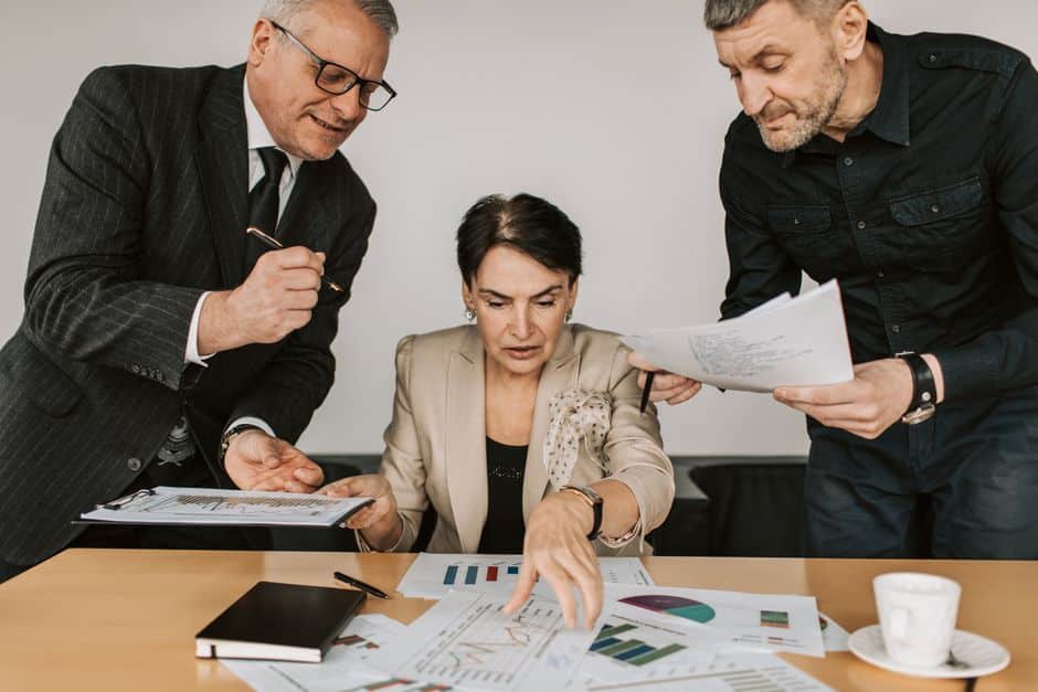 Three professionals in a meeting review charts and documents spread on a table, with one person pointing at a graph.